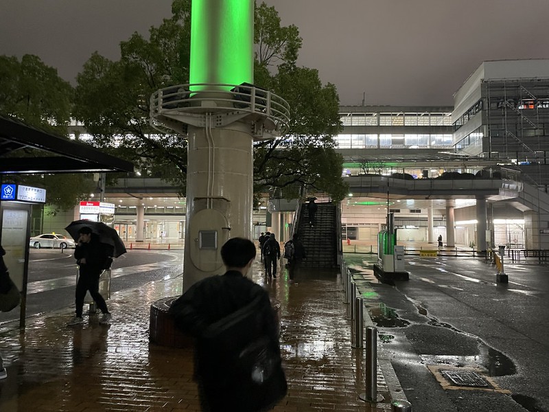 A few people hurry through the nighttime rain outside brightly lit Kokura Station, Japan