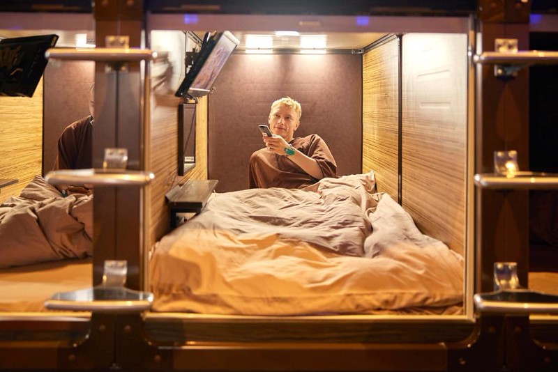 A man sits up in bed changing channel on the TV in the Capsule Hotel Anshin Oyado Premier Tokyo Shinjuku Station