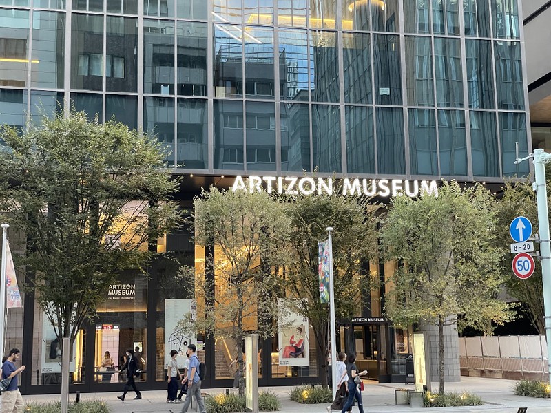 People walk past the exterior to the Artisan Museum in Tokyo, Japan, with the name of the museum in white across the top of the large entrance area and trees outside People walk past the exterior to the Artisan Museum in Tokyo, Japan, with the name of the museum in white across the top of the large entrance area and trees outside