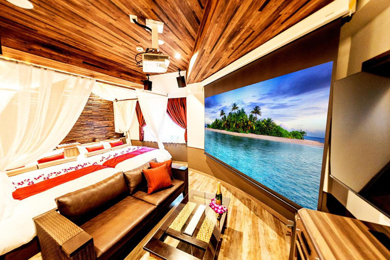 Twin guest room in the BaliAn Resort Forest Ikebukuro, Tokyo, Japan, with two beds, sofa and coffee table, and large projection screen, panelled with dark wood