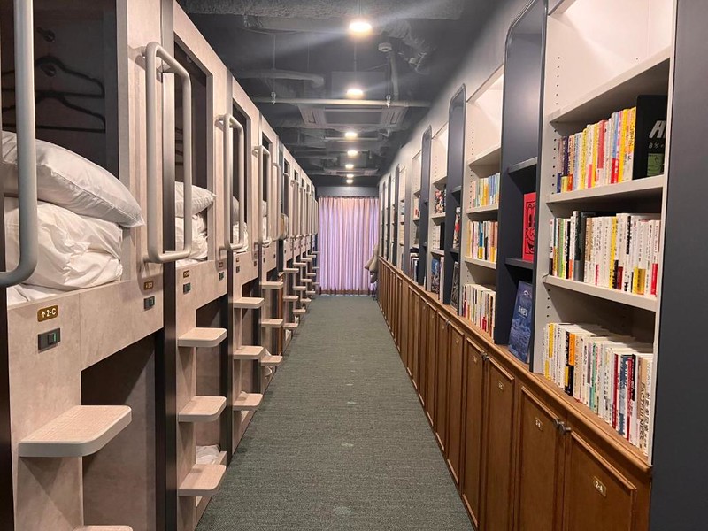 A corridor of double-decker sleeping capsules on the left with shelves of books for guests to read on the right in the Book Tea Bed Shibuya capsule hotel, Tokyo
