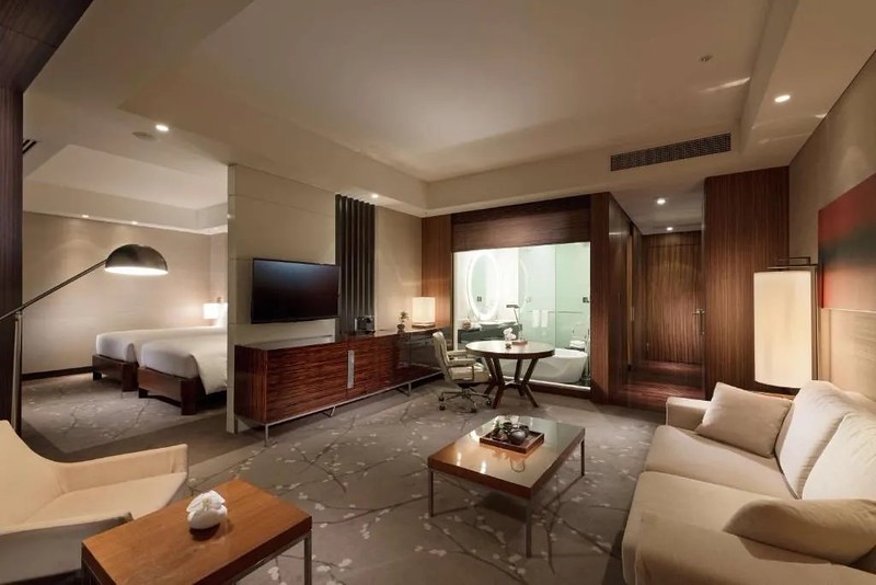 Twin guest suite in the Conrad Tokyo hotel with large living area with sofa and armchair, cherry blossom carpet, and glass unit with bath and large illuminated mirror, as well as separate room with two beds