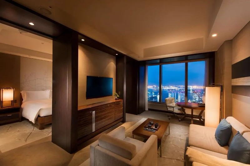 Living area of a Conrad Tokyo hotel suite with sofa and armchair around a wall-mounted TV, separate sleeping area beyond, and dining area beside the window overlooking the nighttime bay