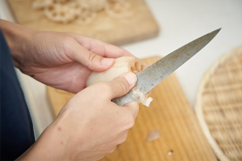 Two hands hold a distinctive white renkon or lotus root while peeling with a long blade. Chopping boards are seen on the table beneath