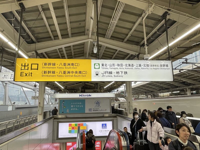 View of a Tokyo train station platform with down escalators and informational signs for exits in yellow and other train lines in white