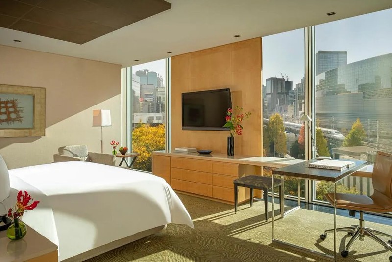 Clean, contemporary double guest room in the Four Seasons Hotel Tokyo, Japan, with L-shaped desk with work chair and stool, fitted chest of drawers, armchair, and bright view of the shinkansen train lines