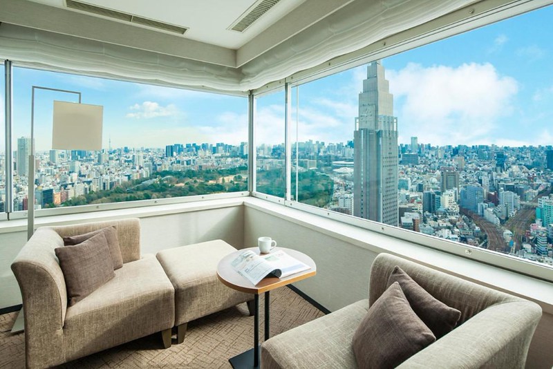 Corner guest room on the upper floors of the Hotel Century Southern Tower, Shinjuku, Tokyo, Japan, with chaise longue and armchair, looking out on the city skyscrapers and large park area