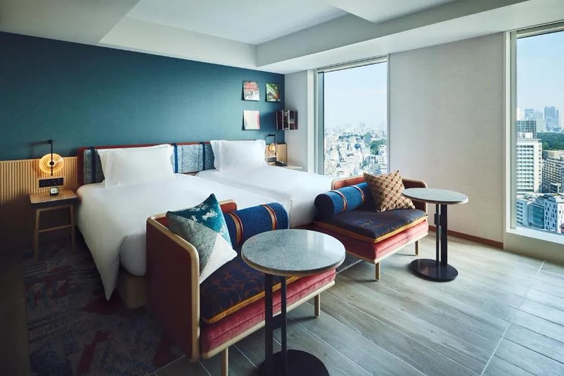 Twin guest room in the Hotel Indigo Tokyo Shibuya, Japan, with joined twin beds with two sofa chairs at the foot and bleached wooden floors, as well as city views