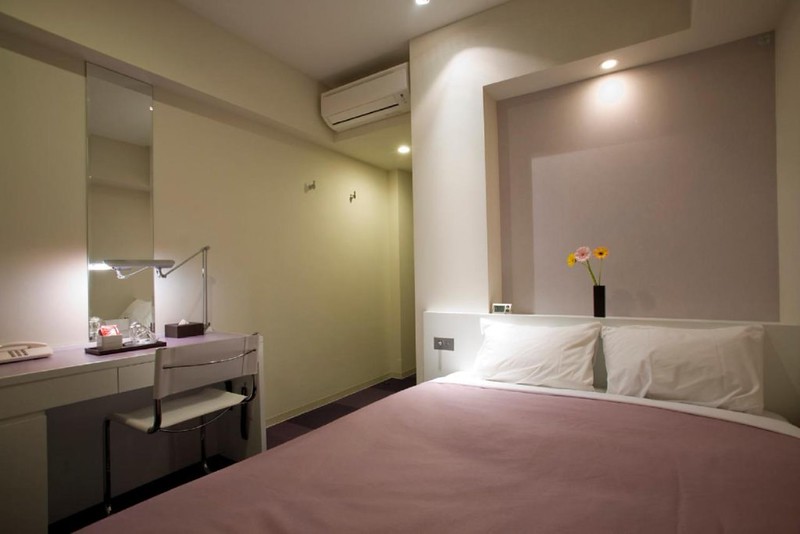Simple guest room in the Hotel Mid in Meguro Ekimae, Tokyo, Japan, with pink bed and matching desk with anglepoise lamp, and shelf above bed with small vase of flowers