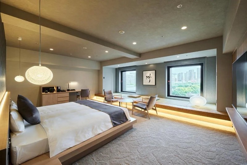 Large guest room in the Hotel Villa Fontaine Shiodome-Tokyo with large mattress on a built-in wooden platform, over patterned rug, and with desk area, twin easy chairs and coffee table, and platform beside windows overlooking city greenery