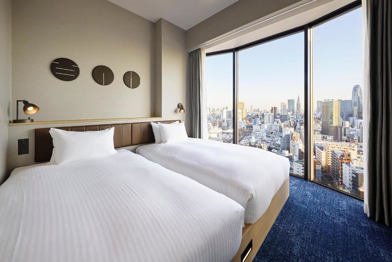 Twin beds in a guest room in the Hundred Stay Tokyo Shinjuku hotel, with city view from the curved windows