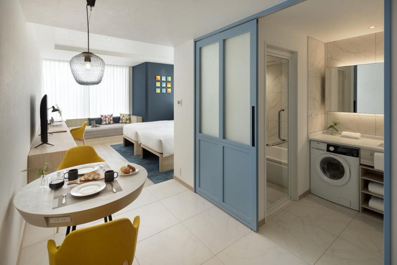 Long view of a small Hyatt House Tokyo Shibuya apartment with twin beds, window sofa, two-person dining table, and bathroom with washing machine and bathtub