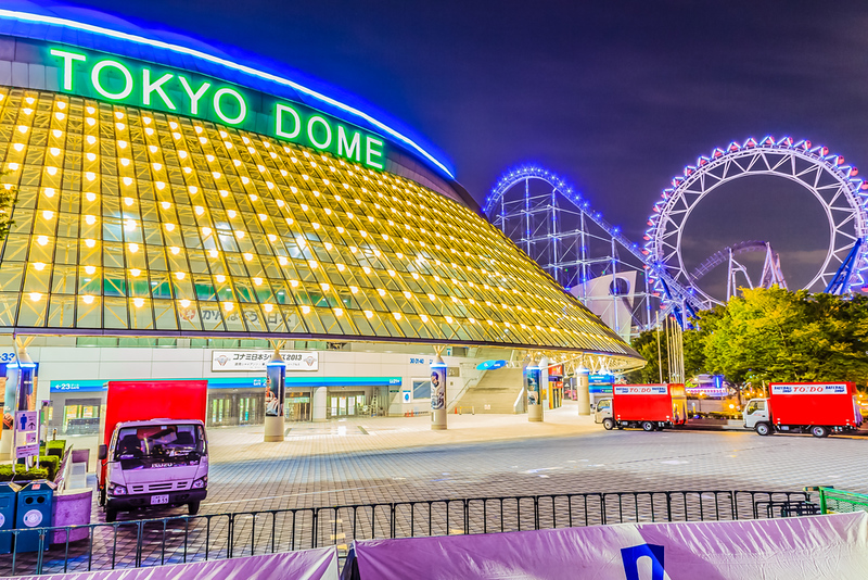 The yellow illuminated roof of Tokyo Dome, with fairground rides in the background, against the evening sky, and a number of small red trucks outside
