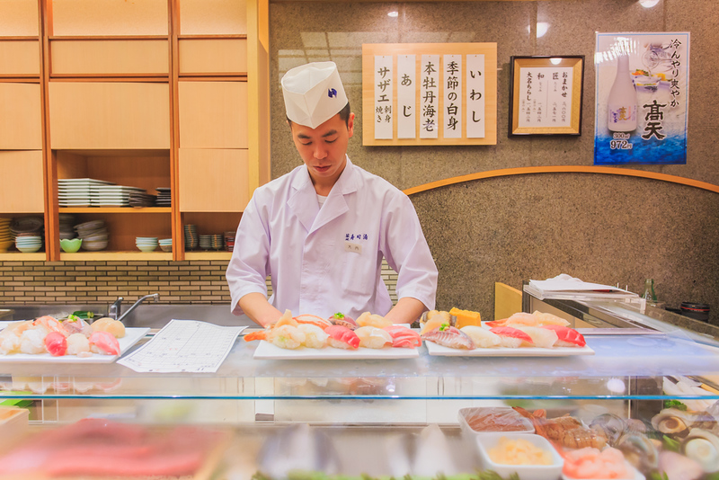 A chef wearing branded whites in Tsukiji, Tokyo, Japan, prepares plates of large sushi pieces from behind a restaurant counter