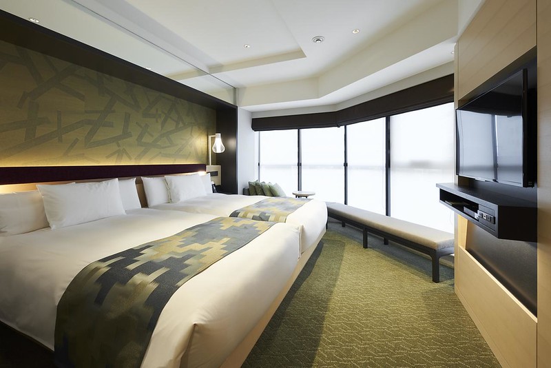 Angular twin guest room in the Hotel 1899 Tokyo, with two large beds, contemporary interior stylings with green patterns, wall-mounted TV unit and windows spread across two walls