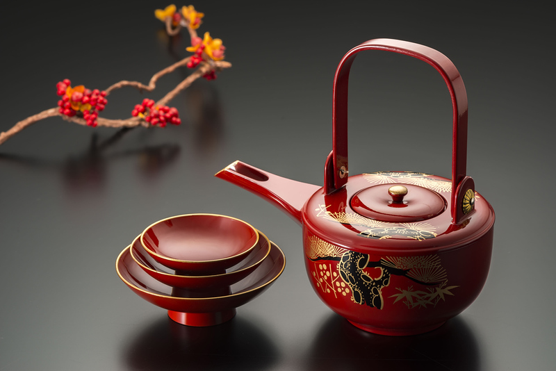 A fine Japanese red lacquerware tea pot next to a stack of small vessels for drinking tea on a black counter next to a twig with red berries