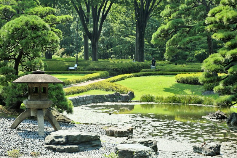 A single visitor wanders through the vibrant green trees, hedges, moss, and grass, broken up with a pond and stone garden in Tokyo’s Imperial Palace Garden, Japan