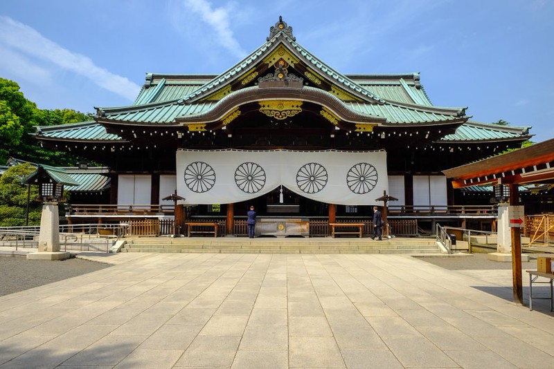 Two separate visitors stand before the traditional green roofs of the Yasukuni shinto shrine in Tokyo, Japan, at the end of a wide expanse of stone slabs, on a bright day