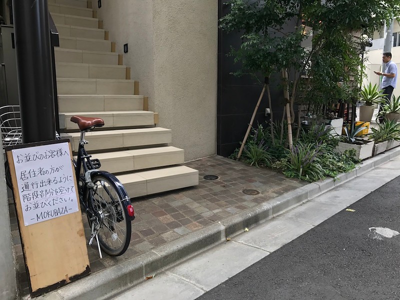 The handwritten sign reads “To customers who are queuing: please leave a space in the queue in front of the staircase so that the residents can pass through.”