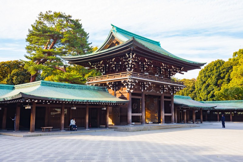 The traditional green curved roof and intricate wooden framework of the Meiji Shrine in Shibuya, Tokyo, Japan, surrounded by green trees with empty stone slabs in the foreground and a single visitor leaving the building