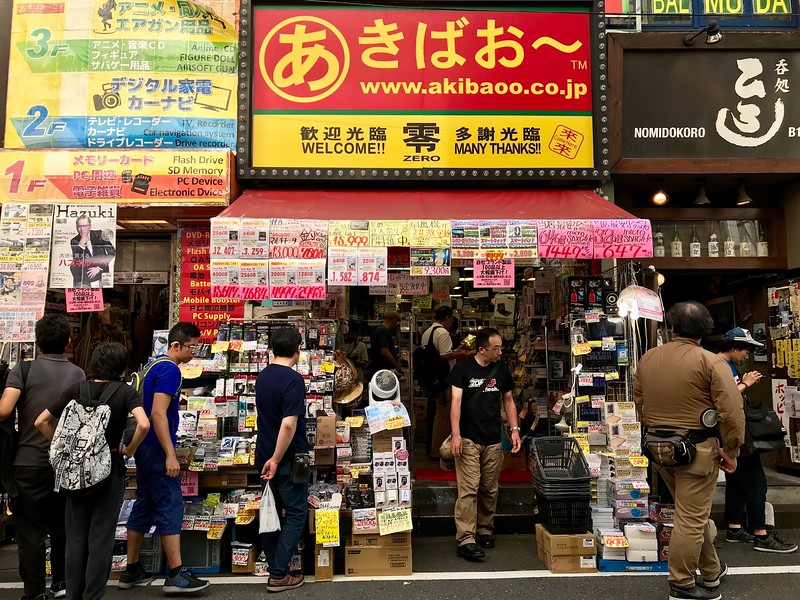 Customers browse outside a small, busy shop in Tokyo’s Akihabara Electric Town, with small electronics accessories on display and small signs displaying prices