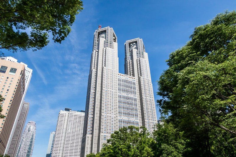 View through the trees, looking up at the towers of the Tokyo Metropolitan Government building against the blue, clear sky