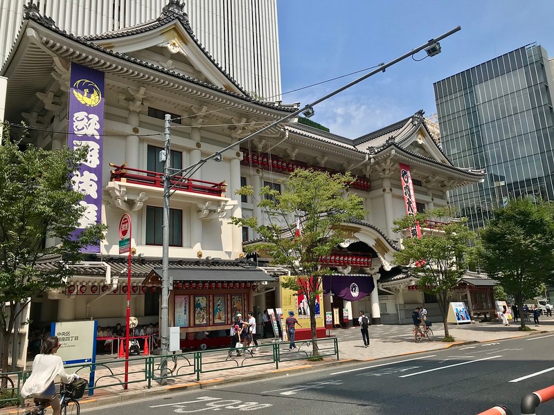 View from the street of the traditional architectural stylings of the main entrance to the Kabukiza Theatre in Tokyo, Japan, with a handful of pedestrians on the pavement outside and modern buildings in the background