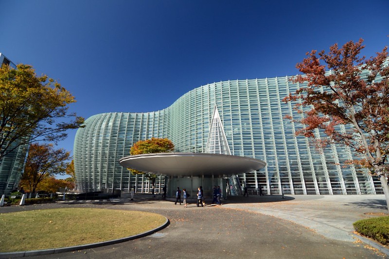 A handful of visitors stand outside the curved glass exterior of National Art Center, Tokyo, Japan, on a bright, clear day