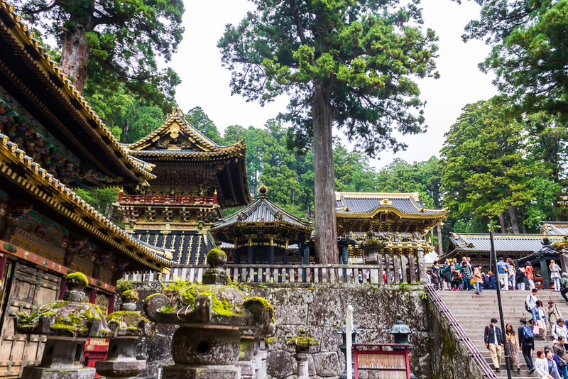 Visitors walk down the stone steps from Tosho-gu Shrine at Nikko, Japan, with mossy sculptures, ornate temple buildings, and tall trees