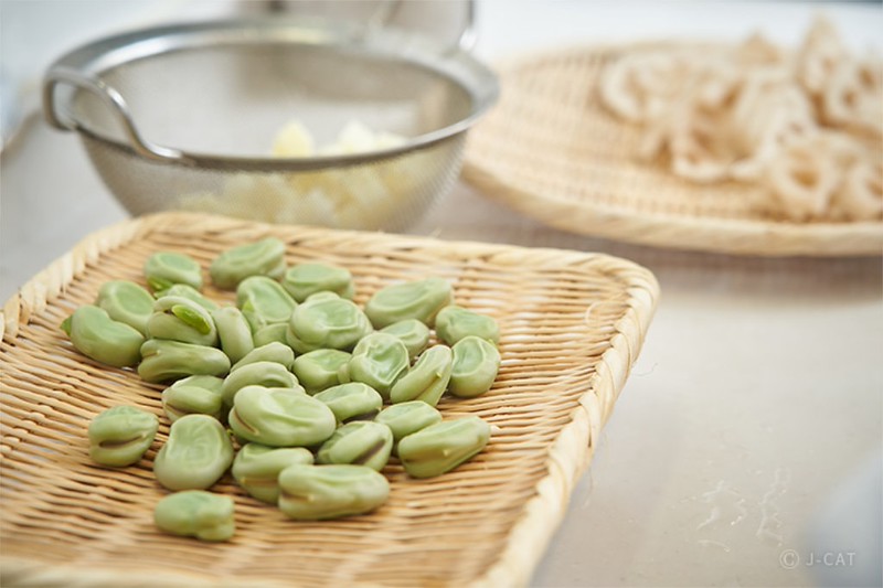 Two wicker plates – one holding broad beans, the other holding lotus roots – sit on a table, next to a metal colander containing another out-of-focus vegetable