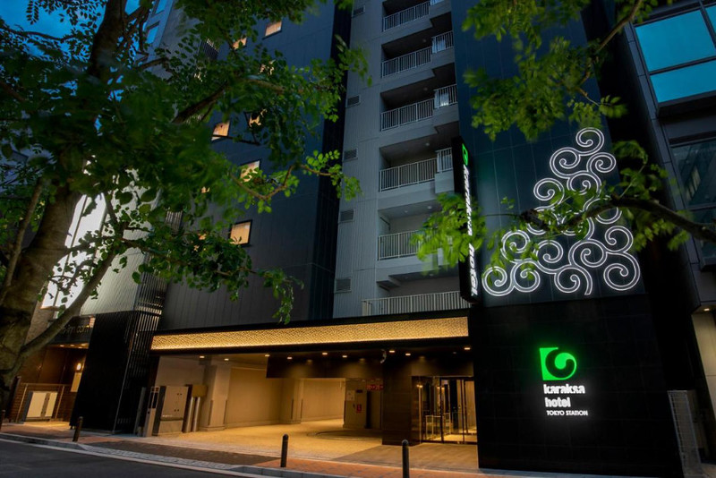 Early evening view of the Karaska Hotel Tokyo Station exterior with illuminated porch, hotel logo and name, and trees on the sidewalk