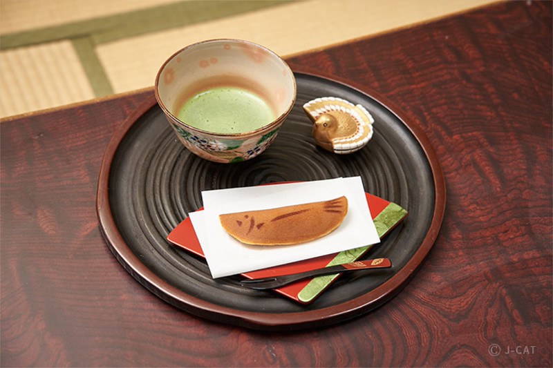 A small cup of matcha tea sits on a distinctive brown plate with a small bird ornament, carefully arranged fork and a fish-shaped pancake-style sweet