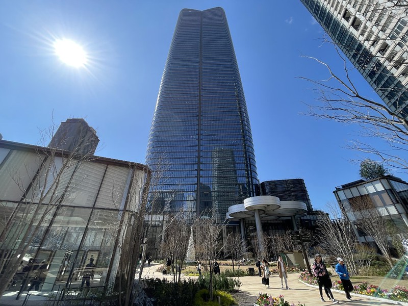 A view from ground level of the Mori JP Tower rising above the Azabudai Hills complex with a glaring sun in the blue sky