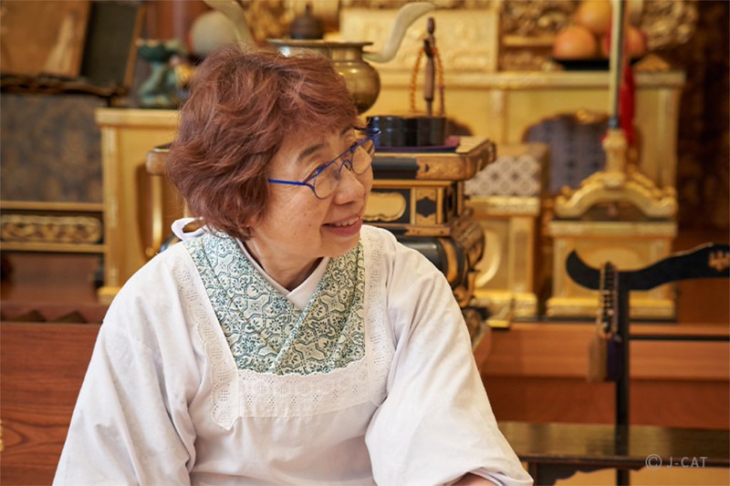 An older Japanese lady named Mr. Asano wearing glasses and cooking robes smiles and looks to an unseen interlocutor
