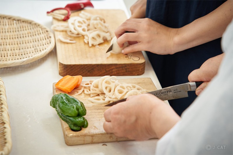 Two sets of hands slice distinctly shaped lotus roots onto thick wooden chopping boards with long cooking knives. A red vegetable peeler and other vegetables are also visible