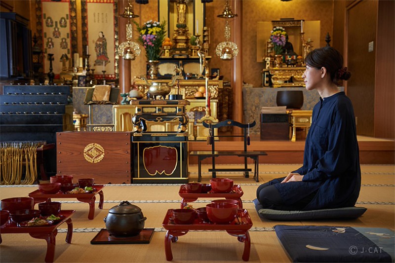 A young woman, dressed in blue, kneels on a cushion in front of red laquered tray tables and a temple altar, ready to eat shojin ryori dishes