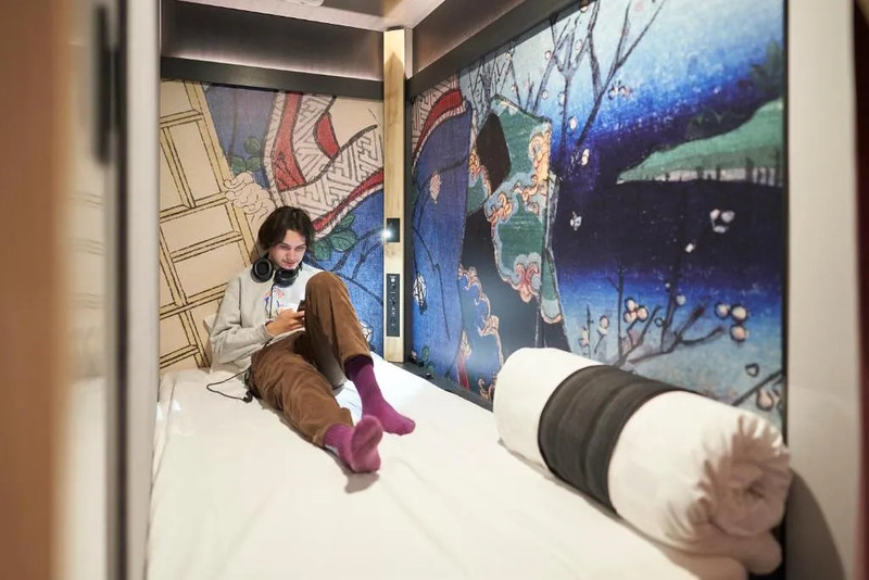 A guest wearing headphones looks on his phone as he sits barefoot on the bed in a brightly-decorated capsule in the Resol Poshtel Tokyo Asakusa hotel