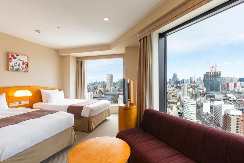 Twin corner guest room in the Shinagawa Prince Hotel, Tokyo, Japan, with two wide beds, and sofa against the wide red window length sofa and view overlooking the city