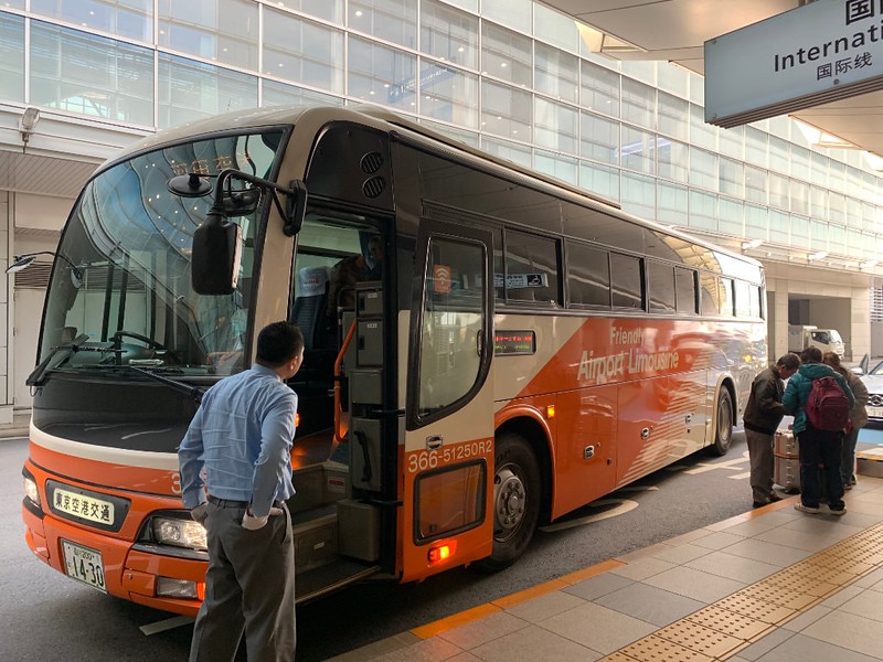 Passengers stand next to an orange and white airport limousine bus outside Haneda International, Tokyo