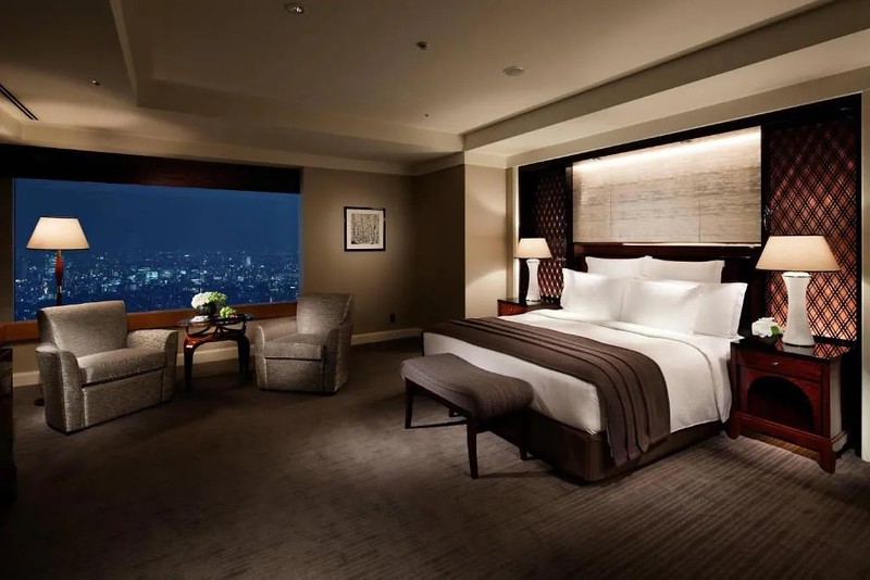 Large double guest room in The Ritz-Carlton Tokyo hotel, Japan, with bed and twin armchairs, brown striped carpet, and wide windows with nighttime city views