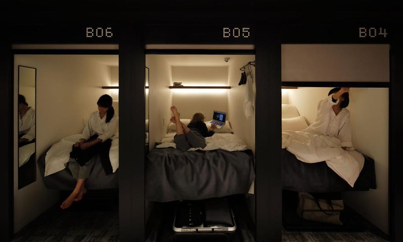 Young women in three sleeping pods in The Millennials Shibuya hotel in Tokyo – one sits on the bed and packs a day bag, one lies on her stomach looking at a laptop, and the other pulls the shade down ready to go to sleep