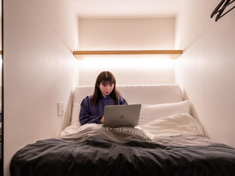 A young Japanese woman sits up in bed with a laptop in an illuminated sleeping pod in The Millennials Shibuya hotel, Tokyo