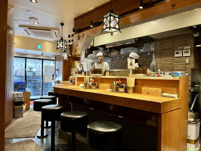 Two members of staff wearing kitchen whites stand behind the counter of a small ramen restaurant in Tokyo