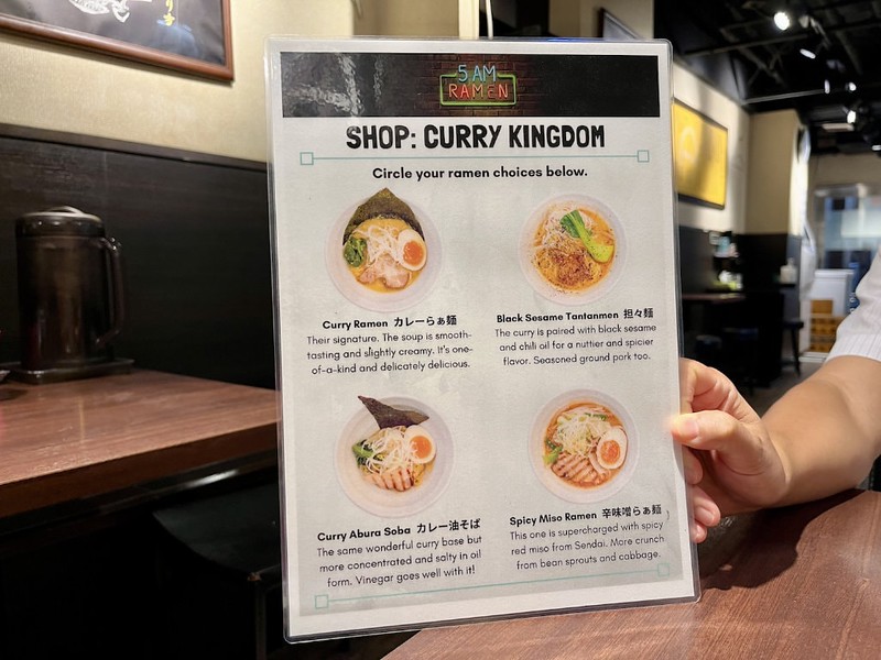 A food tour guide holds up a laminated menu of different ramen options to choose with pictures of each dish and explanations in English of what they contain